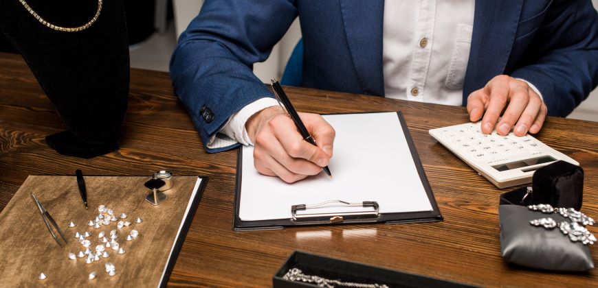 Person writing on clipboard with jewelry and a calculator