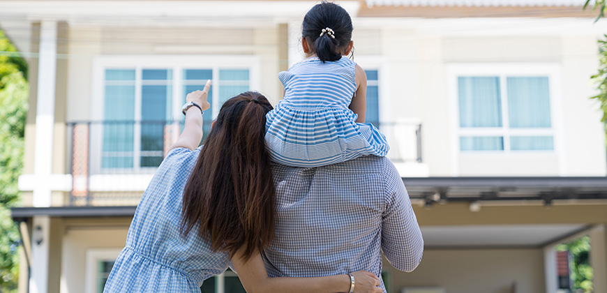happy couple stands and points at house with daughter on their shoulder
