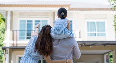 happy couple stands and points at house with daughter on their shoulder