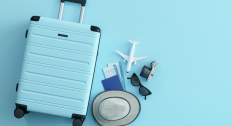 Blue suitcase sitting next to a hat, plane tickets, toy plane, sunglasses, and a camera on top of a blue background