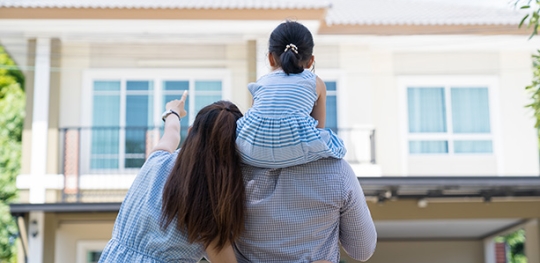 happy couple stands and points at house with daughter on their shoulder